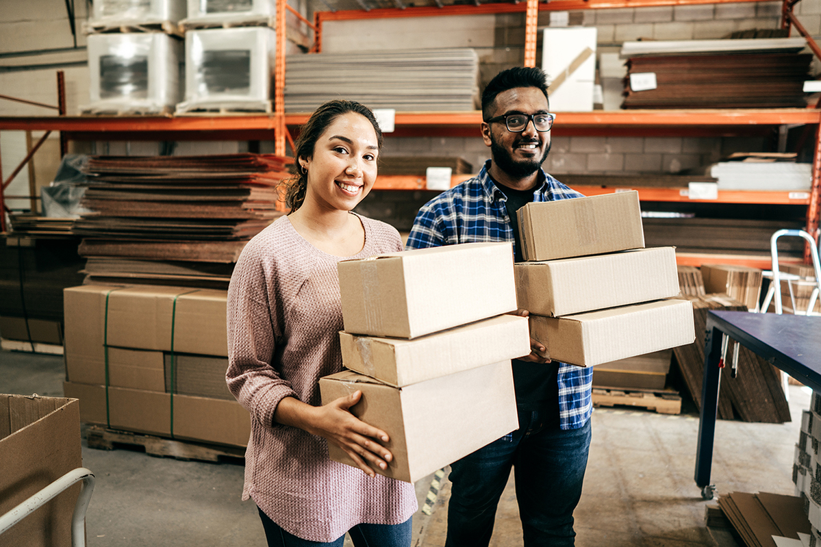 Two smiling employees hold boxes in a warehouse.